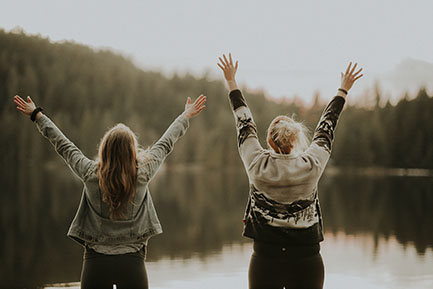 Two women stood with arms stretched out by a lake. Photo by Priscilla Du Preez from Unsplash.